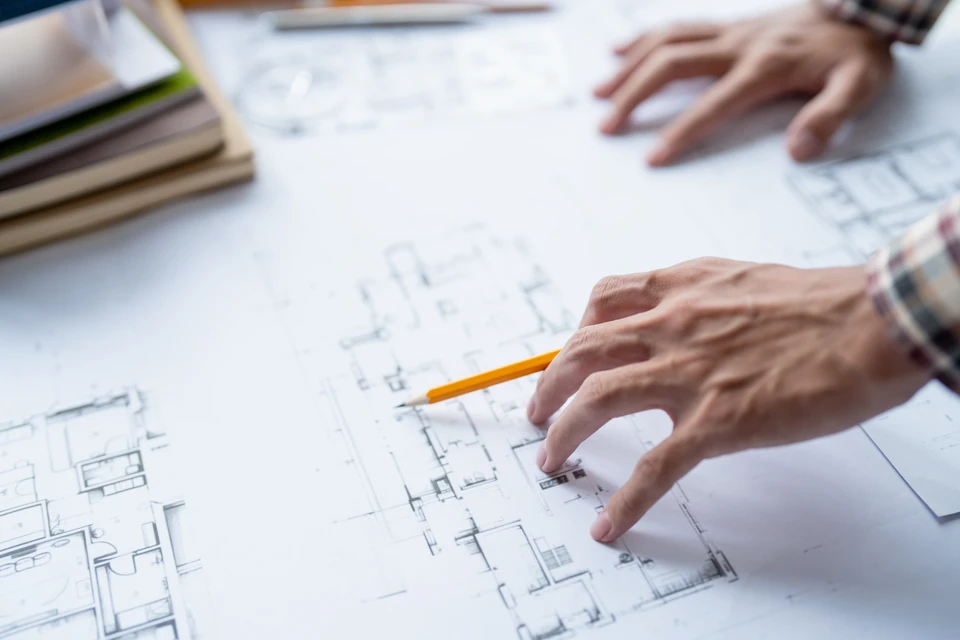 Un chef de projet ou manager de chantier en tenue professionnelle d'entreprise de bâtiment (chemise, gilet de sécurité ou costume), consulter des plans de rénovation sur un chantier multi-corps de métier, avec visible en arrière-plan des artisans et ouvriers travaillant à différentes tâches (menuiserie, plomberie, électricité), symbolisant la coordination et l'orchestration de plusieurs métiers du bâtiment pour une rénovation globale.